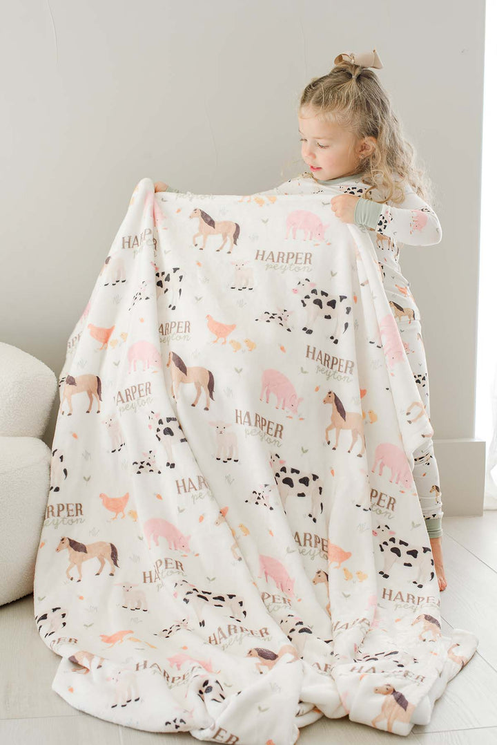 Toddler girl in Caden Lane farm animal pajamas holding up a personalized 'Harper Peyton' farm animal minky blanke