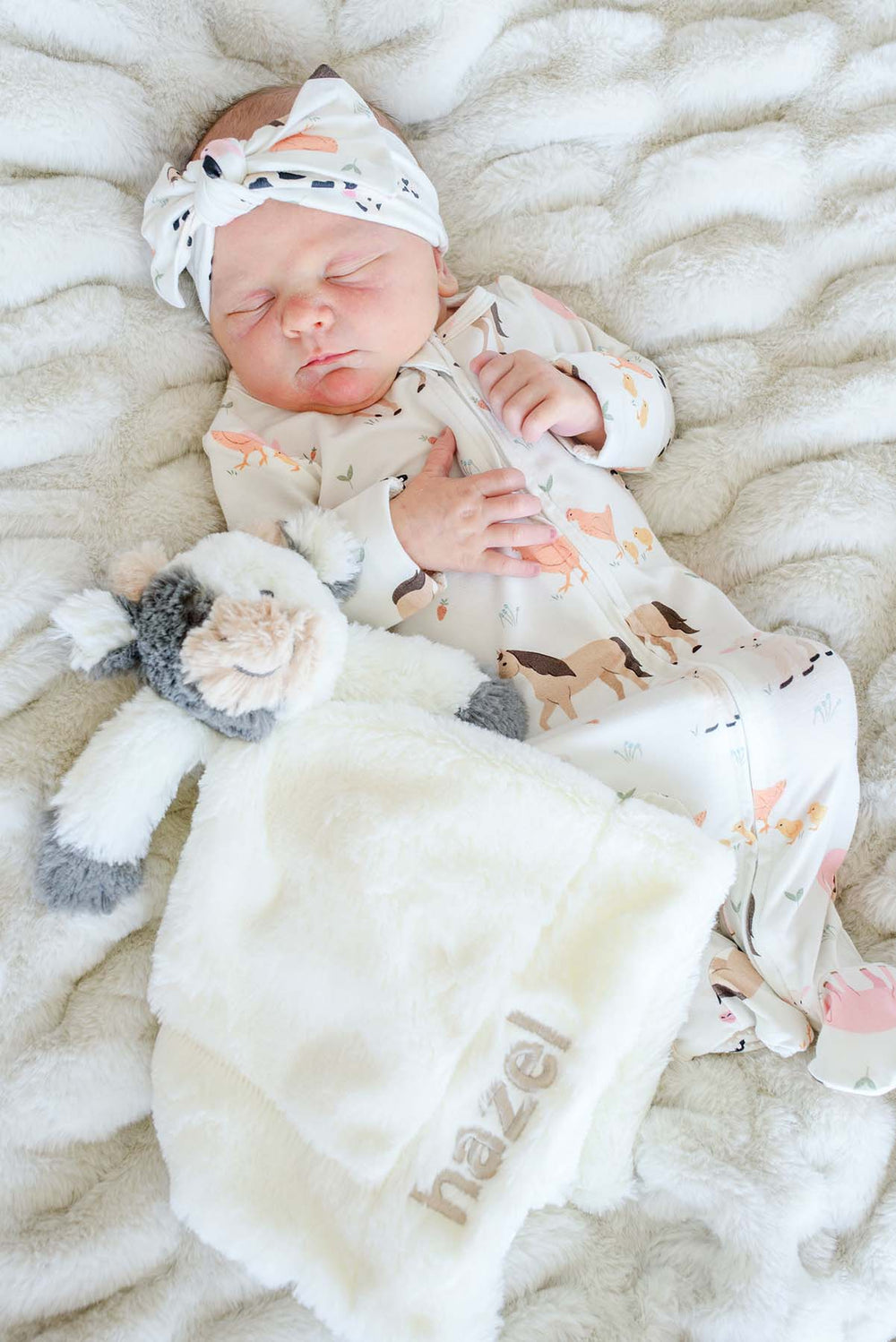 Sleeping newborn in Caden Lane farm animal footie pajamas with matching headband, holding a personalized 'Hazel' cow lovey blanket