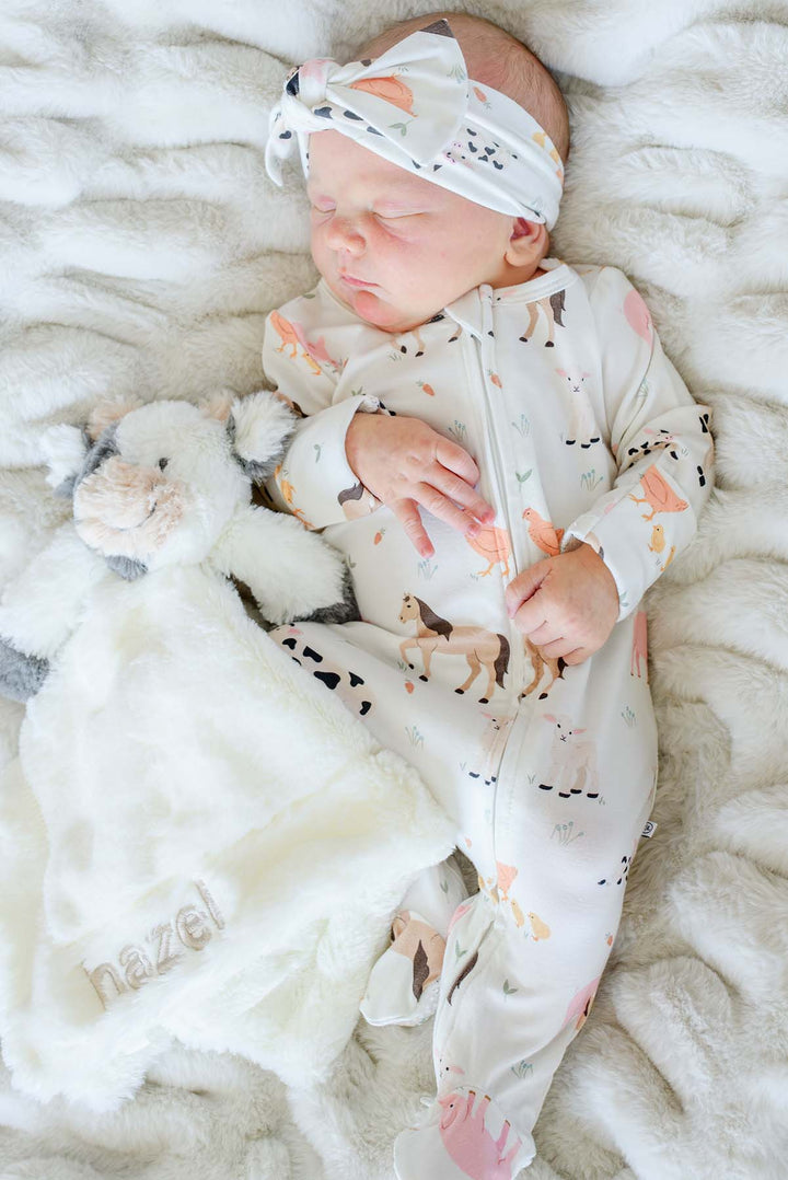 Close-up of sleeping newborn in Caden Lane farm animal footie pajamas and headband, holding a personalized 'Hazel' cow lovey blanket