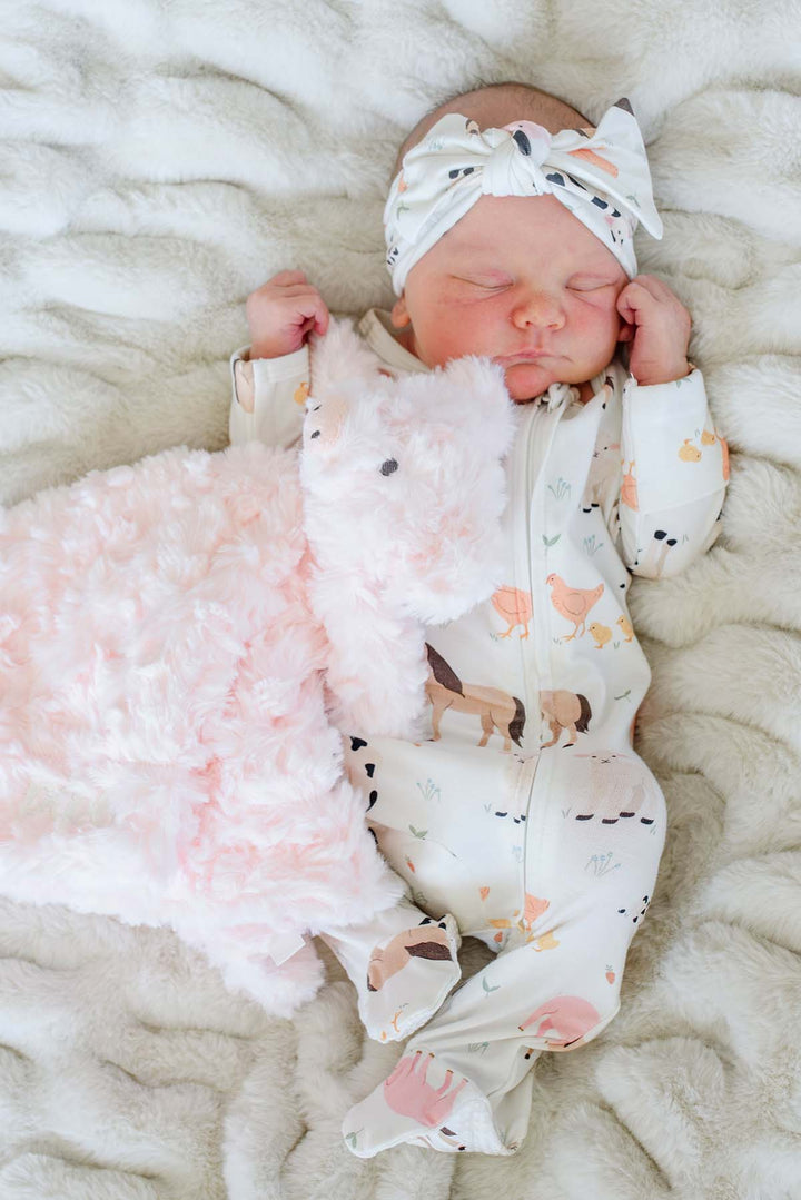 Sleeping newborn in Caden Lane farm animal footie pajamas and matching headband next to a personalized 'Hazel' cow lovey blanket