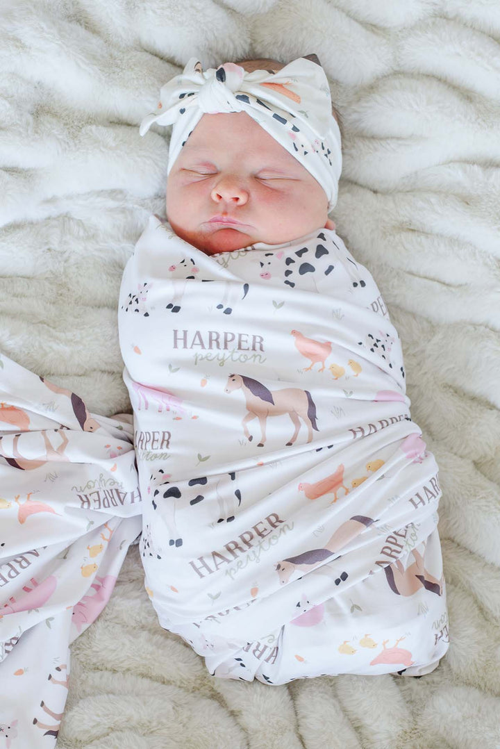 Caden Lane personalized farm animal swaddle blanket with custom name 'Harper Peyton,' shown wrapped around a sleeping newborn with matching knotted headband