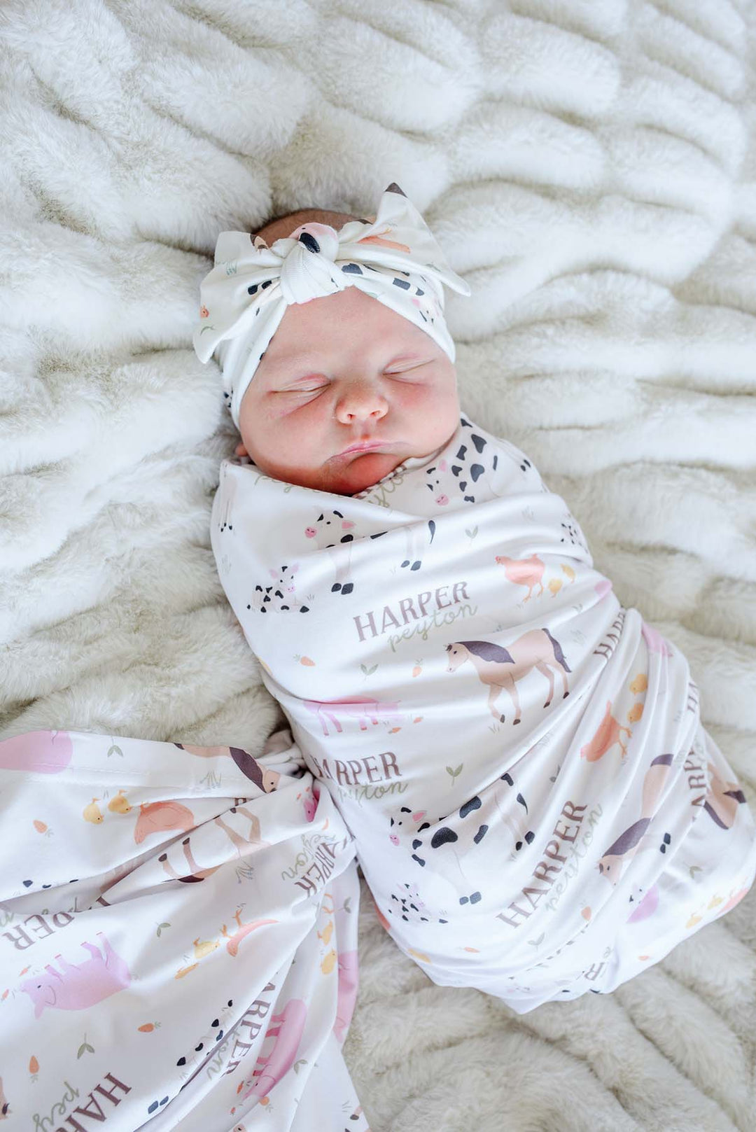 Newborn baby sleeping wrapped in Caden Lane personalized 'Harper Peyton' farm animal swaddle blanket with matching bow headband
