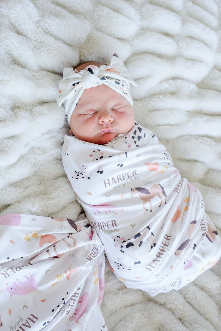 Newborn baby sleeping wrapped in Caden Lane personalized 'Harper Peyton' farm animal swaddle blanket with matching bow headband