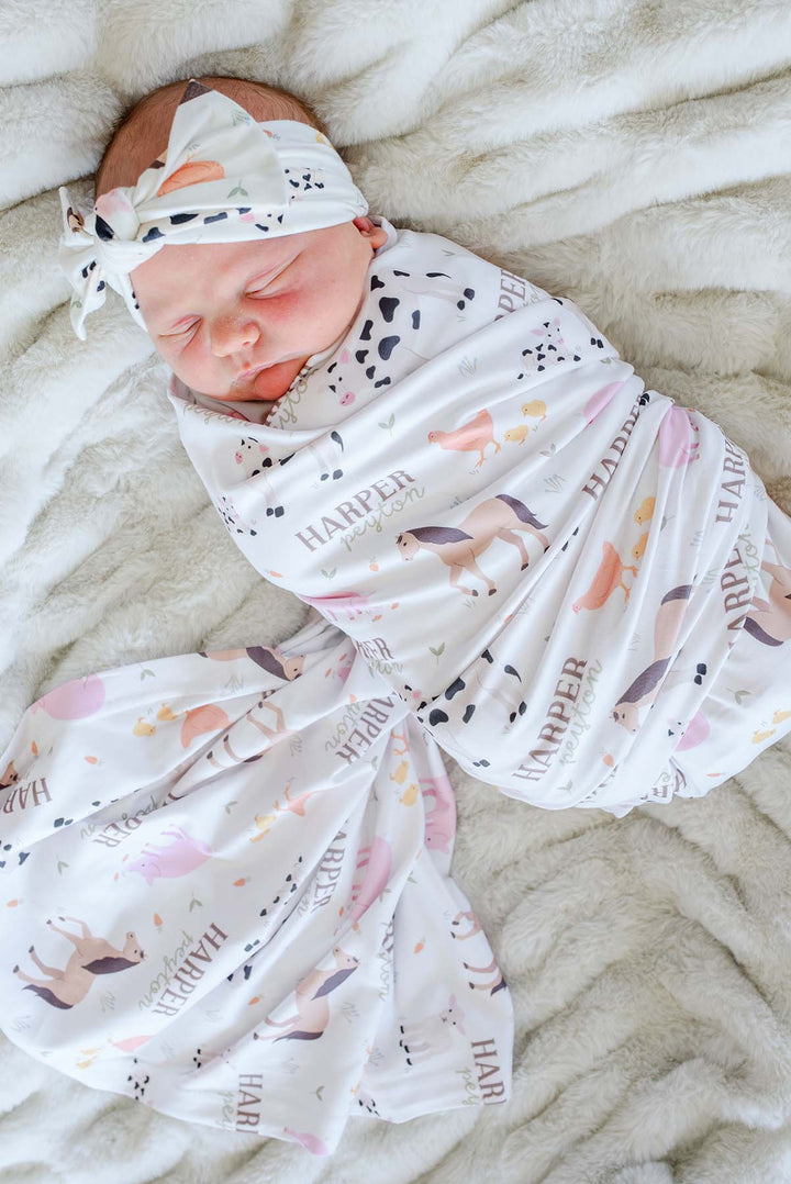 Caden Lane personalized farm animal swaddle blanket and headband set with custom name 'Harper Peyton,' shown swaddling a sleeping newborn