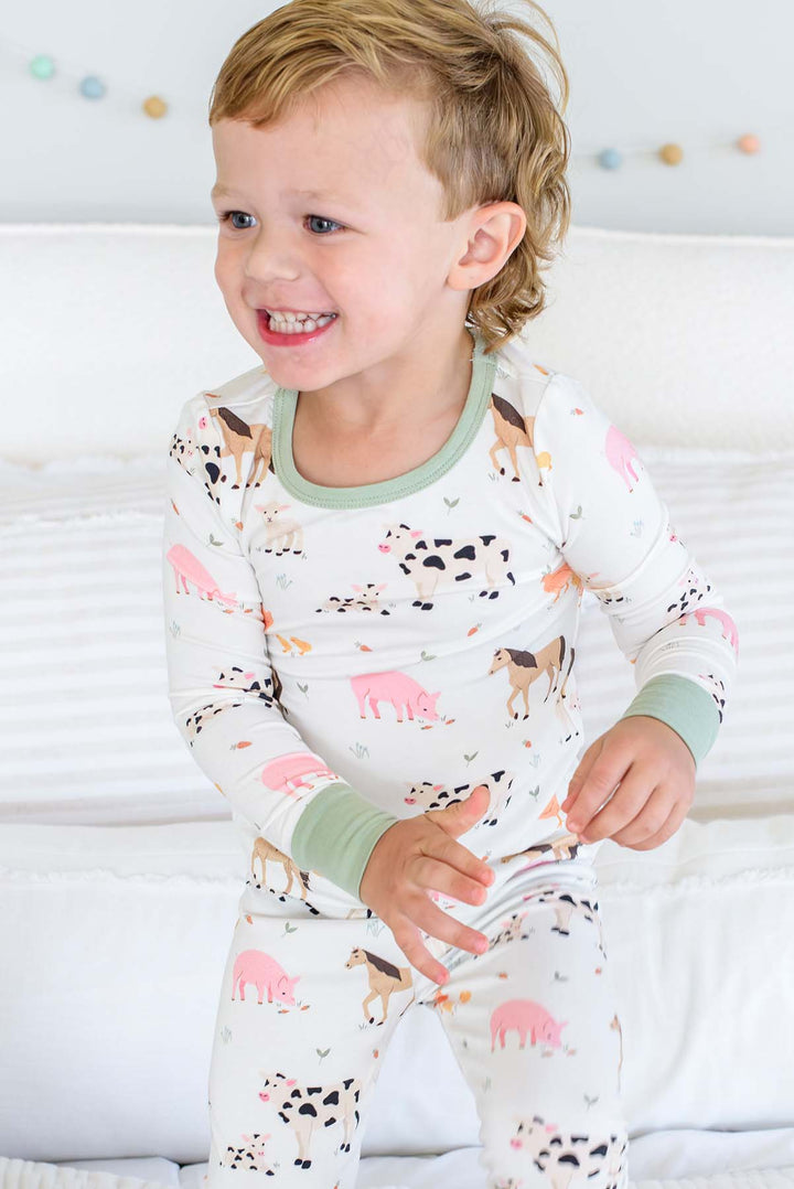 A smiling toddler wearing a Caden Lane long-sleeve pajama set with an animal print, standing on a white bed. The pajamas are white with pink, black, and brown farm animal illustrations and light green trim.
