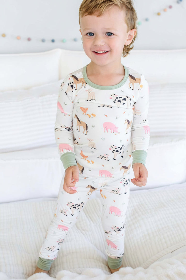 A toddler stands on a white bed wearing a Caden Lane long-sleeve pajama set with an animal print. The pajamas are white with green trim and feature colorful illustrations of farm animals like cows, pigs, horses, and dogs.