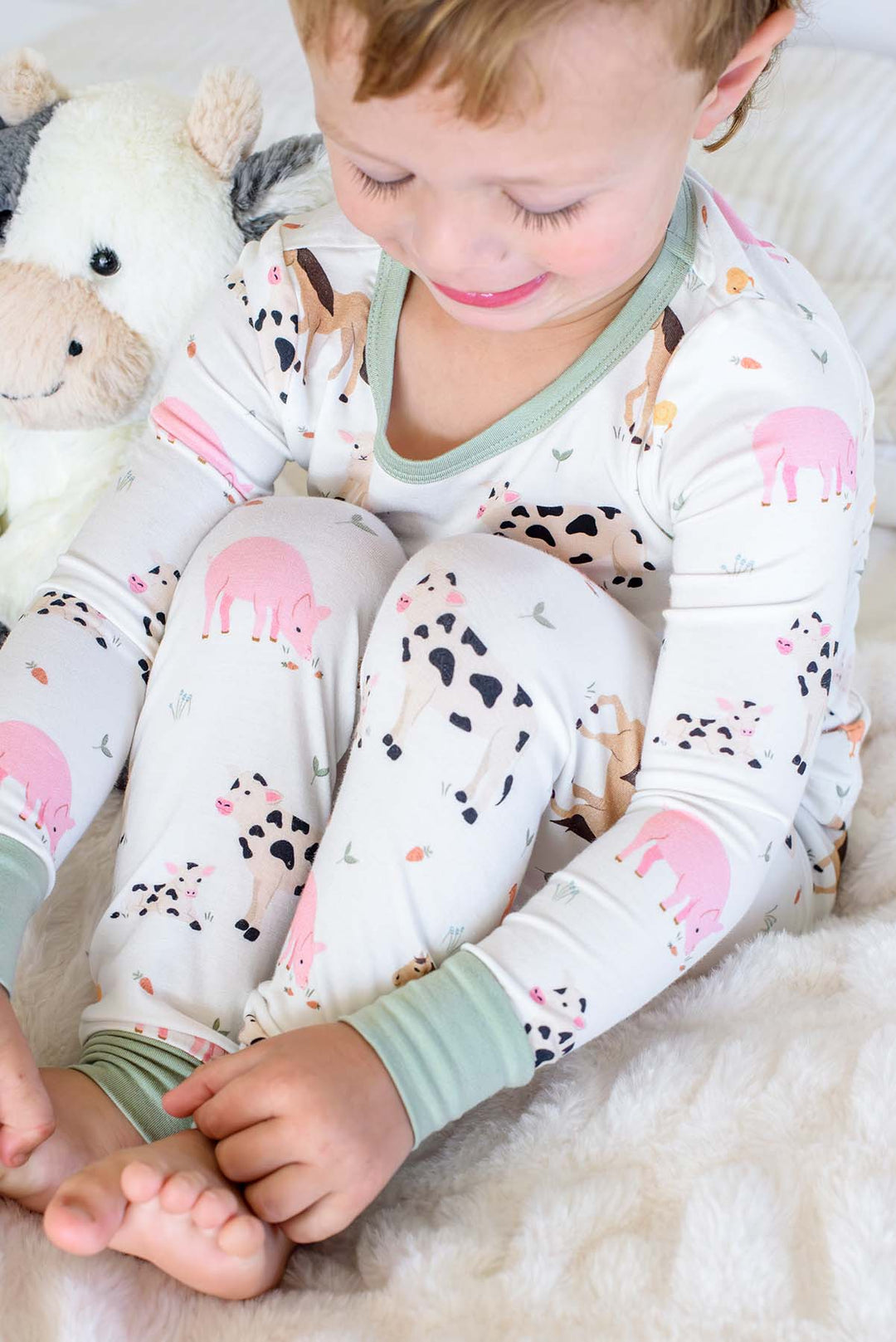 A toddler sitting on a white faux fur blanket wearing a long-sleeve pajama set with a colorful farm animal print. The pajamas are white with pink pigs, black-and-white cows, and light green trim on the collar and cuffs.