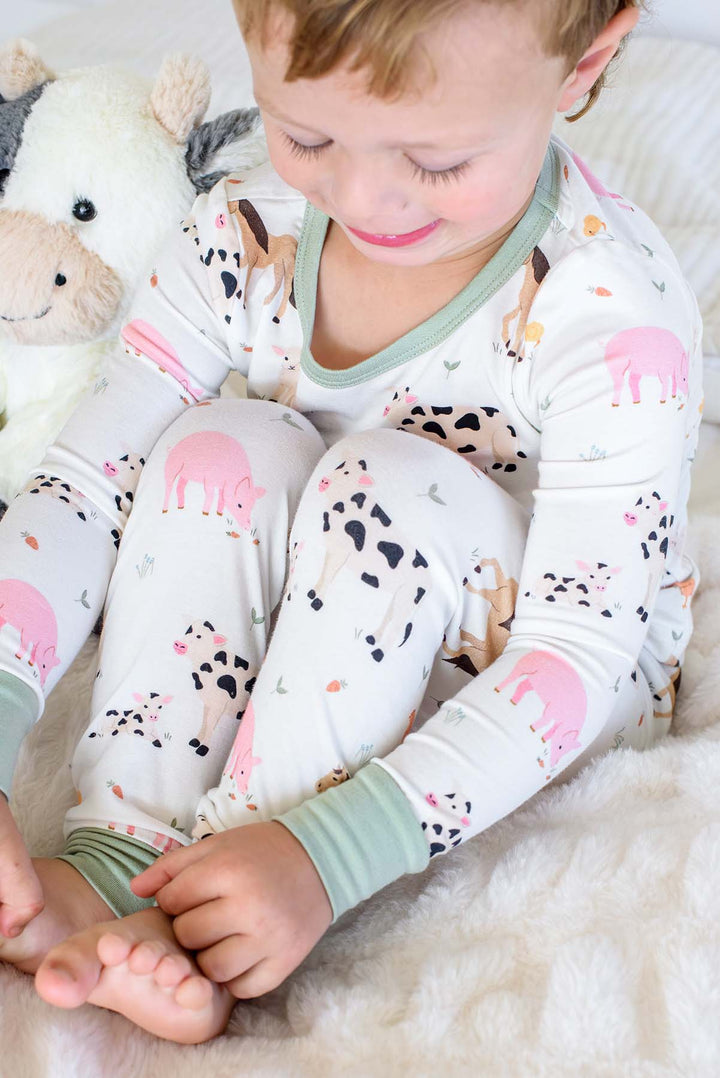 A toddler sitting on a white faux fur blanket wearing a long-sleeve pajama set with a colorful farm animal print. The pajamas are white with pink pigs, black-and-white cows, and light green trim on the collar and cuffs.