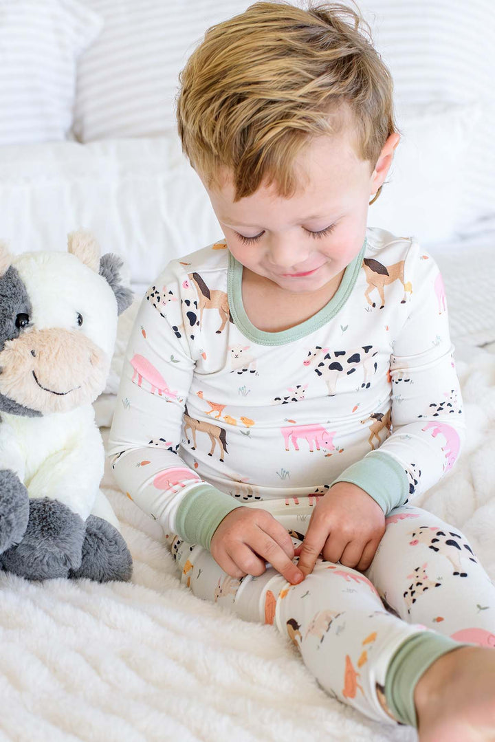 A young child sitting on a white bed wearing a Caden Lane long-sleeve pajama set with a colorful animal print, next to a plush cow toy.