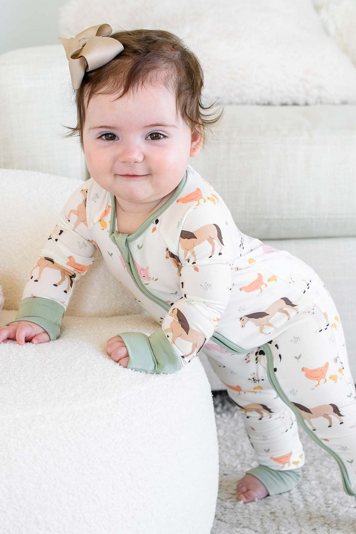 A baby wearing a Caden Lane cream-colored footless pajama romper with a woodland animal print and sage green trim, crawling on a white textured ottoman in a bright room.