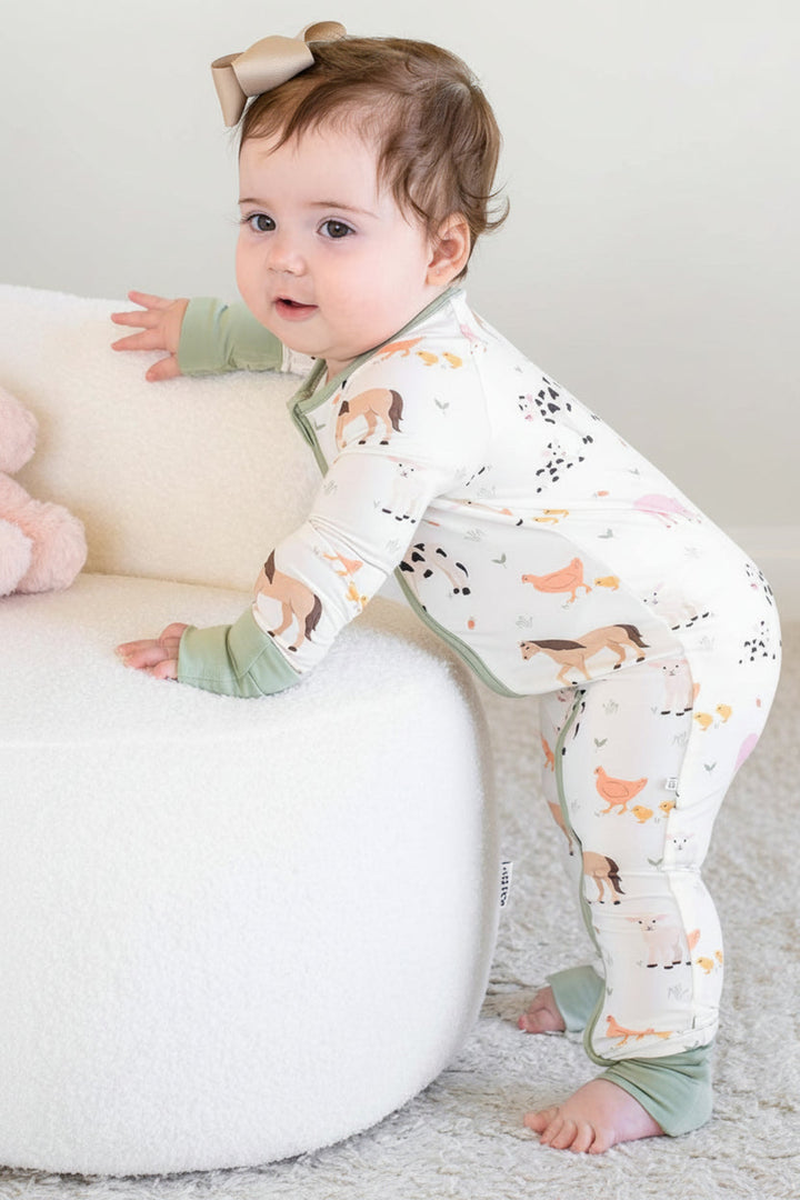 A baby wearing a Caden Lane white long-sleeve footless pajama romper with a colorful fox print and sage green cuffs, standing on a white textured rug next to a white pouf and a pink plush toy.
