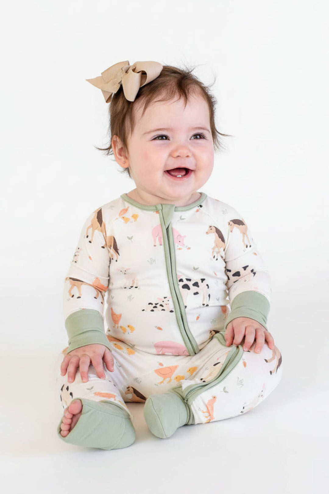 A baby sitting and wearing a Caden Lane cream-colored zip-up romper pajamas with a colorful animal print and sage green trim, along with a matching beige bow headband.