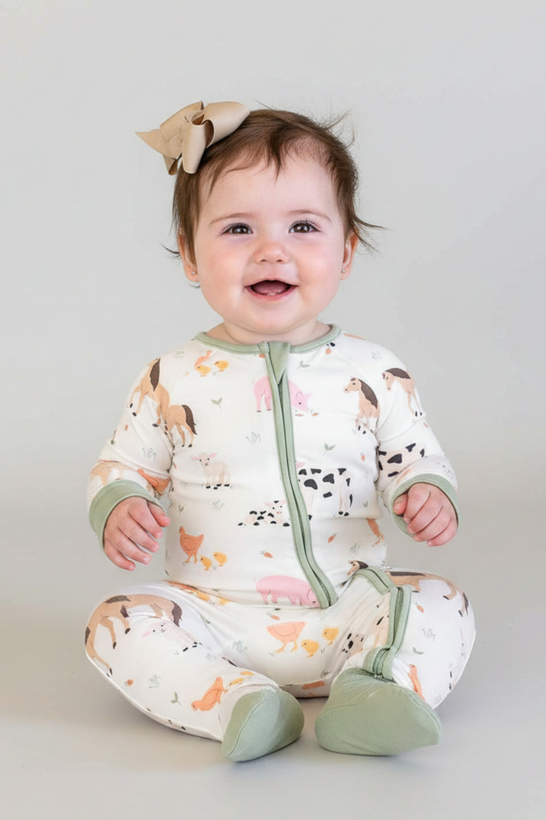 A baby wearing a Caden Lane cream-colored footed sleeper with a green zipper and sage green cuffs, sitting on a white chair. The sleeper has a playful print of various farm animals like cows, pigs, and chickens.