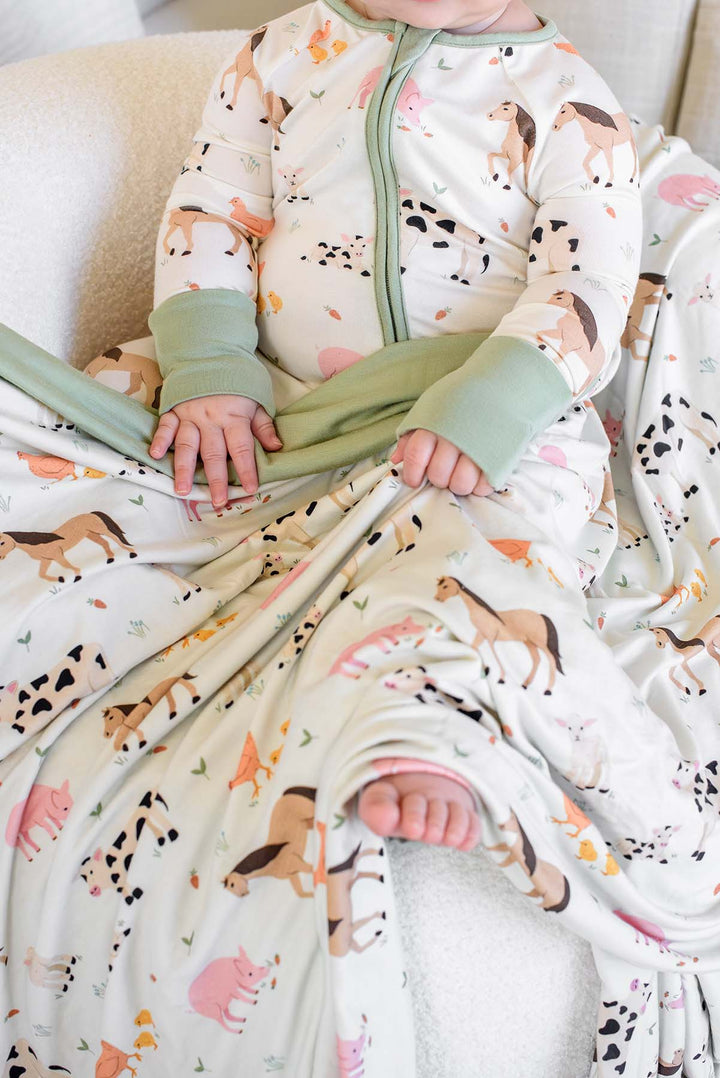 Close-up of baby in Caden Lane farm animal footie pajamas with sage green cuffs, holding matching farm animal blanket