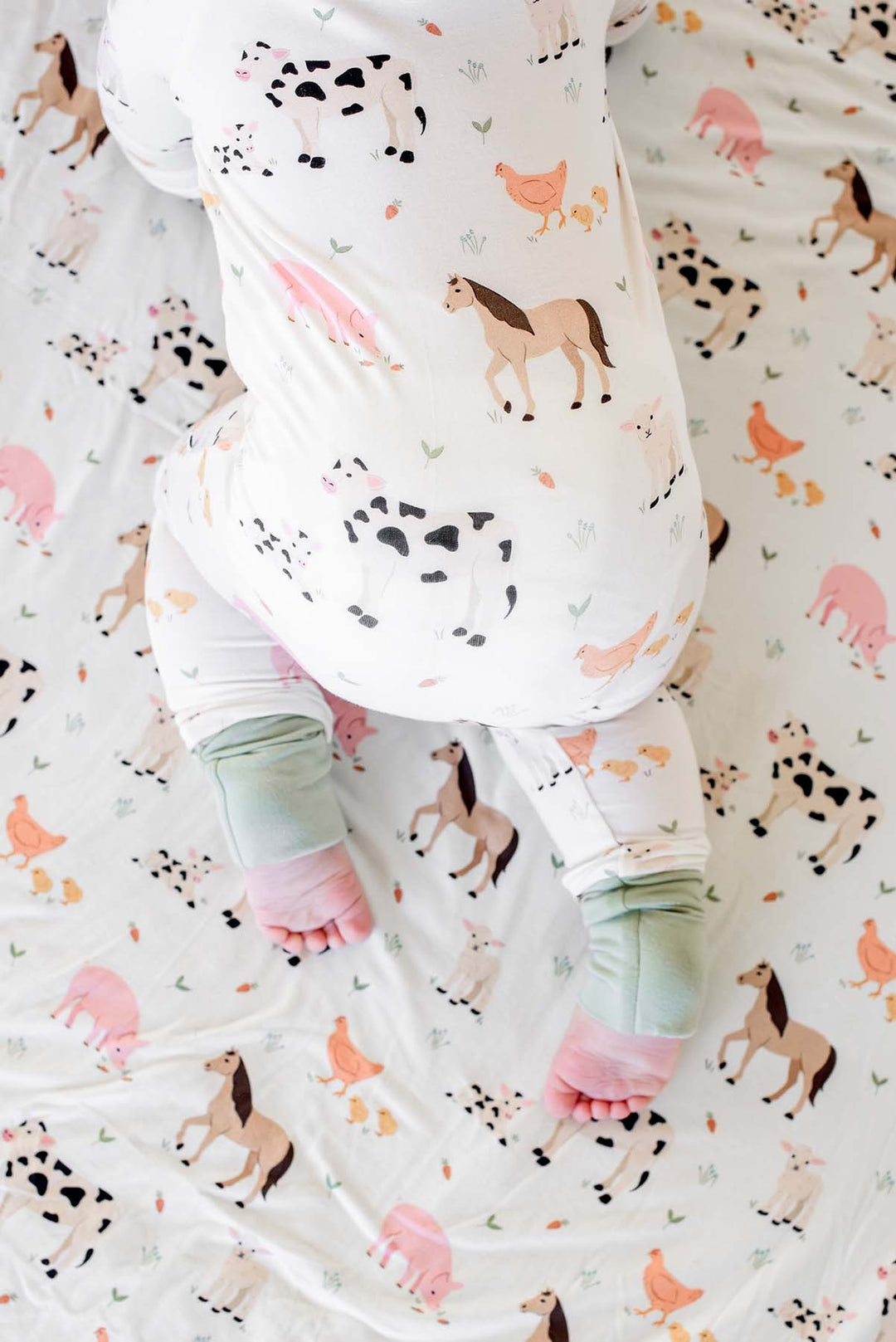 A baby lying on a Caden Lane blanket with a colorful farm animal print, wearing a matching pajamas.