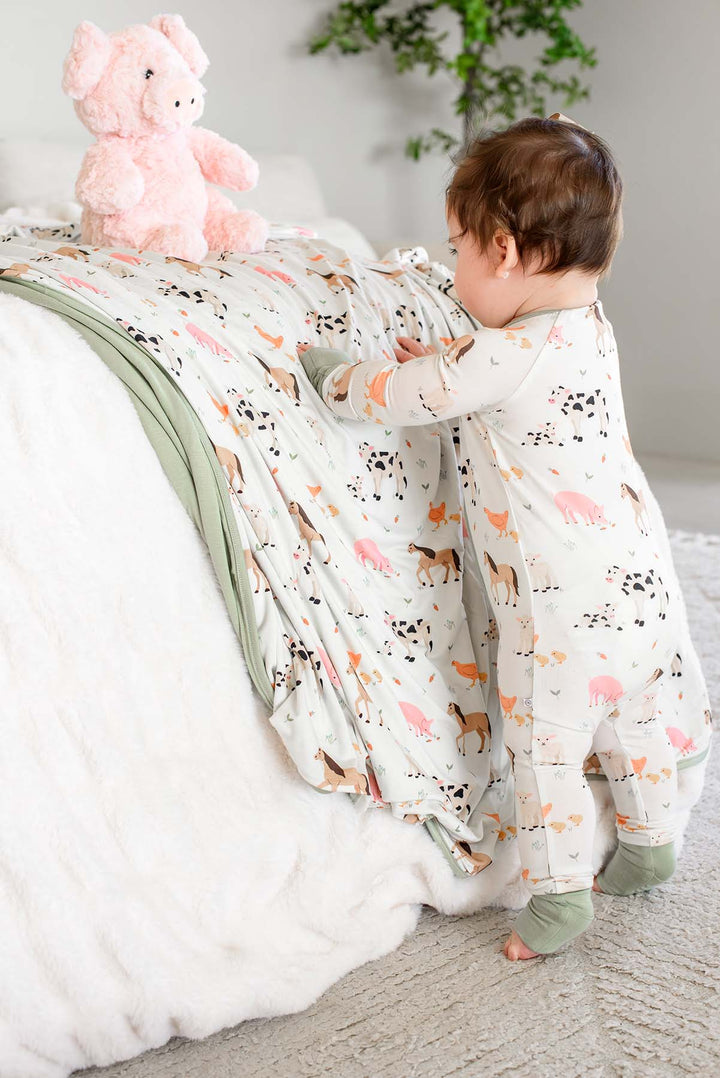 Baby standing at bed in Caden Lane farm animal footie pajamas with sage trim, reaching for stuffed pink pig toy on matching farm animal blanket