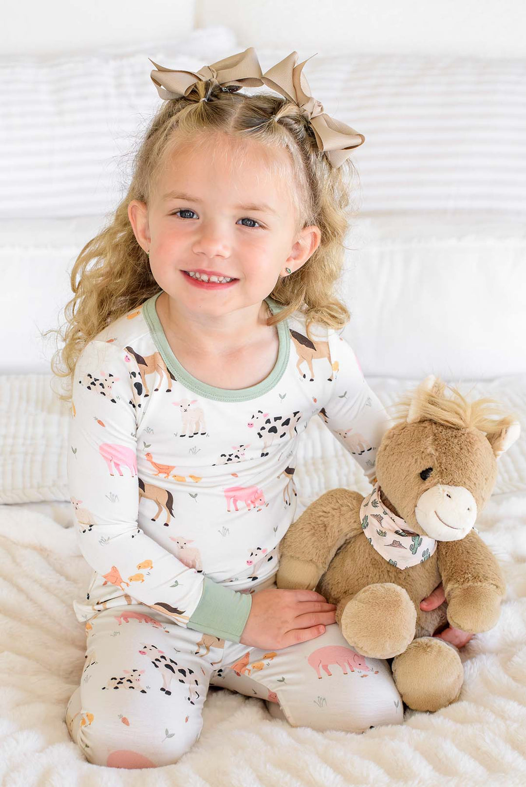 miling toddler girl in Caden Lane farm animal two-piece pajama set with sage trim, sitting with a stuffed horse toy