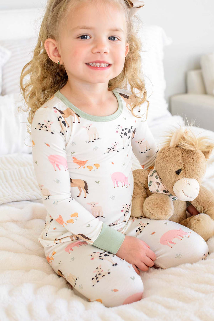 A young girl with blonde curly hair sits on a white bed, wearing a long-sleeve pajama set with a woodland animal print and holding a brown plush toy.