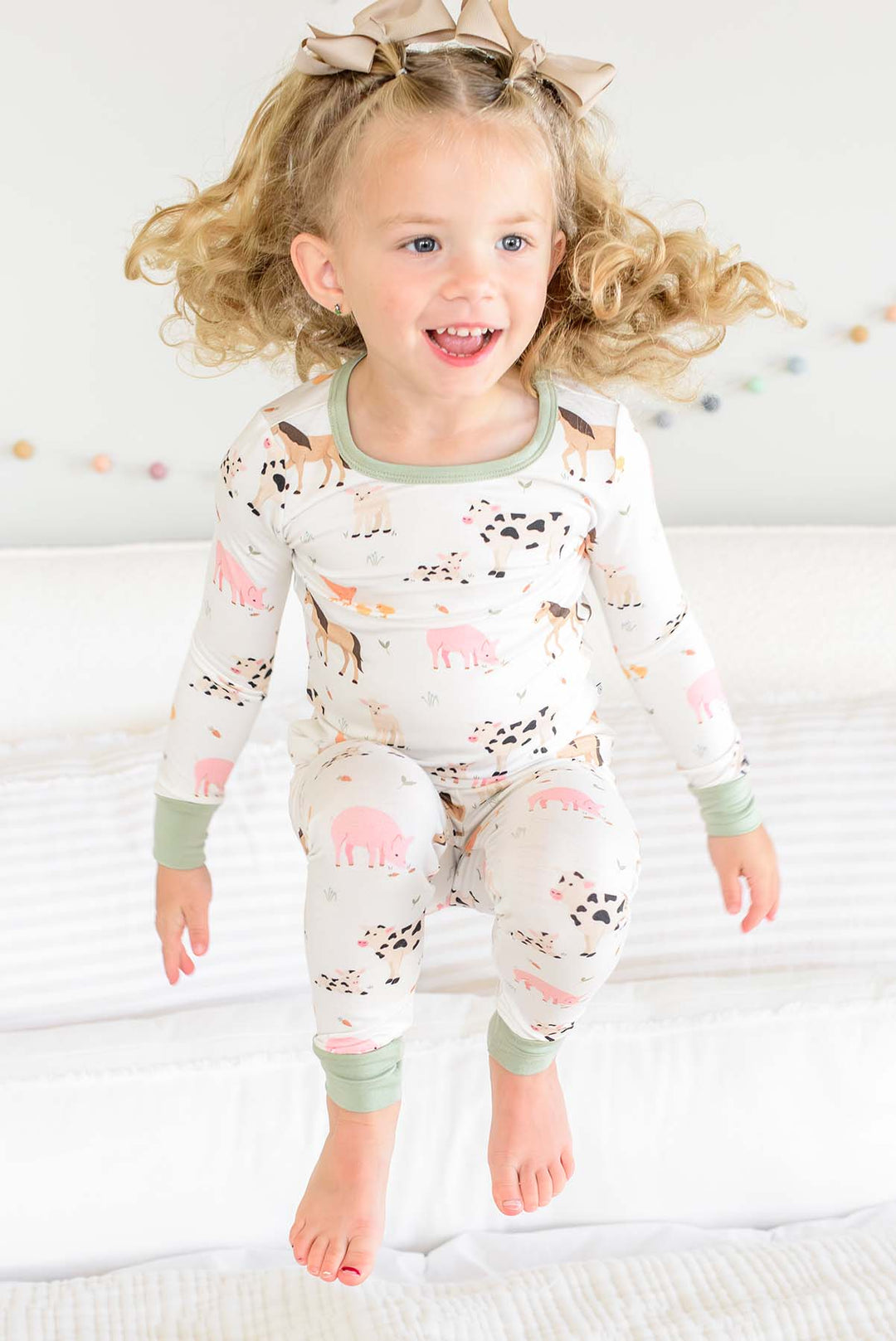 Toddler girl jumping on bed in Caden Lane farm animal two-piece pajama set with sage trim