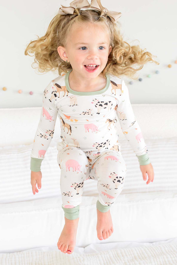 Toddler girl jumping on bed in Caden Lane farm animal two-piece pajama set with sage trim
