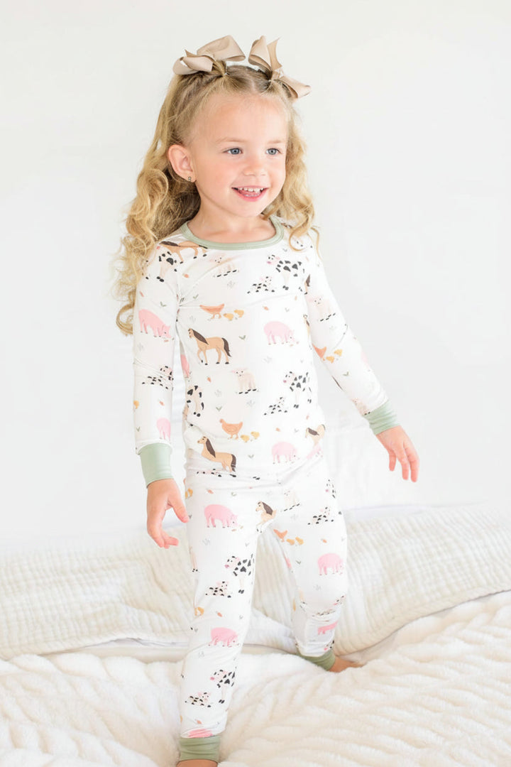A young girl with curly blonde hair wearing a Caden Lane matching two-piece pajama set with a cute animal print, standing on a white quilted bed.