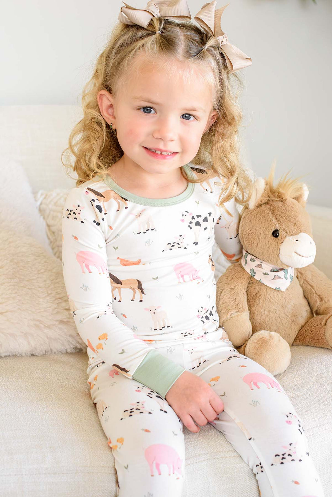A young girl with blonde curly hair wearing a Caden Lanevwhite long-sleeve pajama set with a safari animal print, sitting on a light beige couch next to a brown plush toy horse.