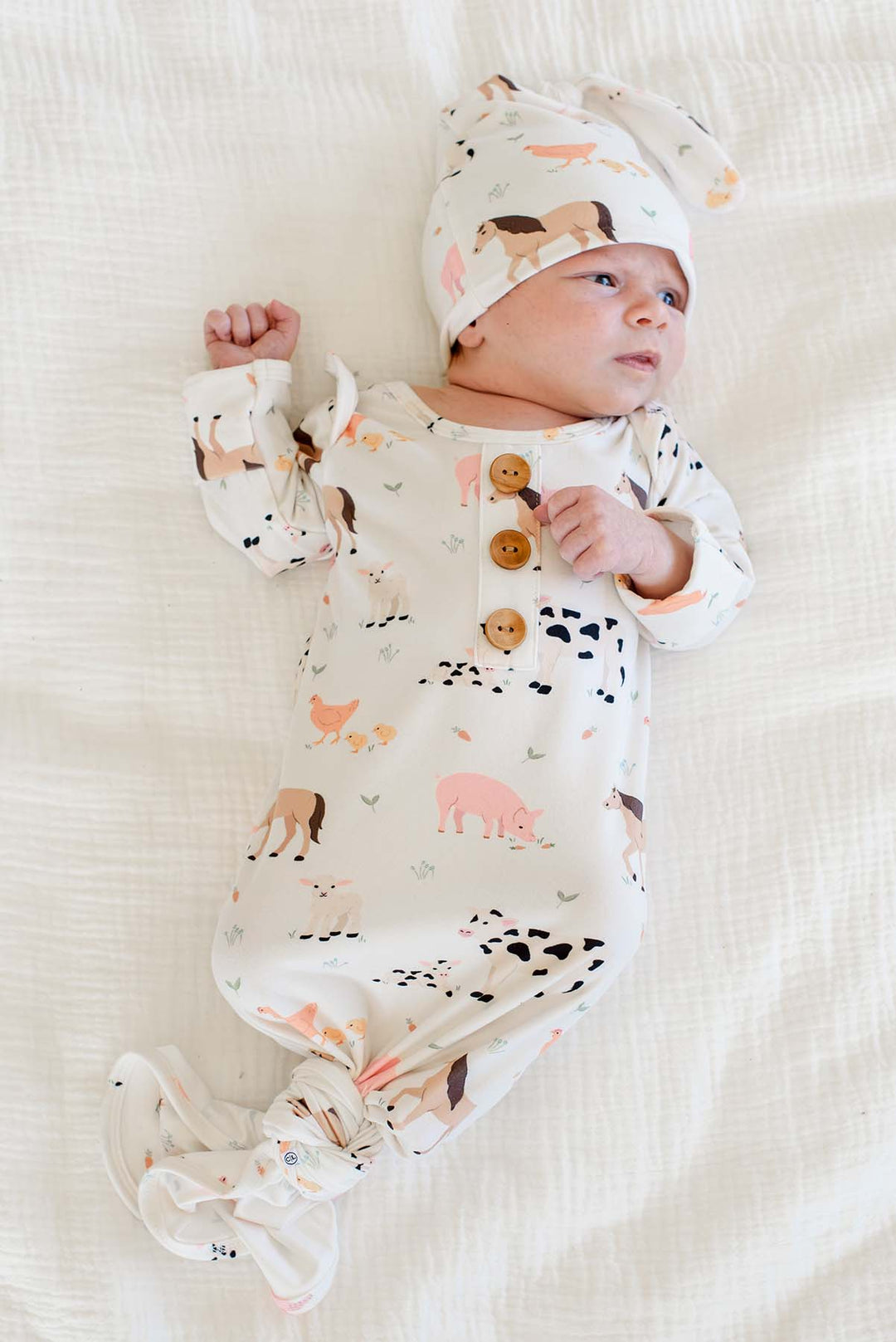 Newborn wearing Caden Lane farm animal knotted gown with wooden buttons and matching topknot hat, lying on white bedding