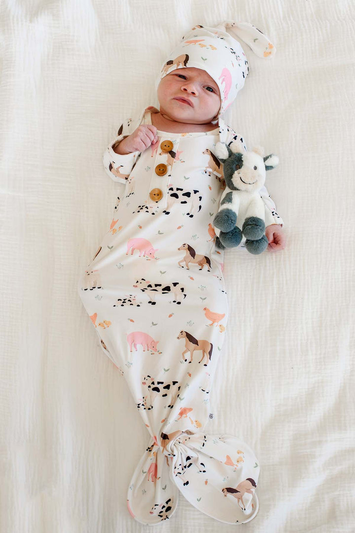 Newborn wearing Caden Lane farm animal knotted gown and matching topknot hat, holding a gray and white cow stuffed toy