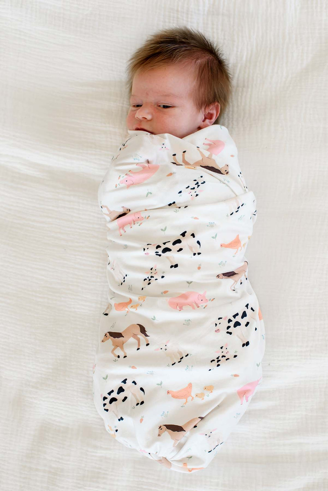 Newborn baby swaddled in Caden Lane farm animal print blanket with pink trim, lying on white bedding