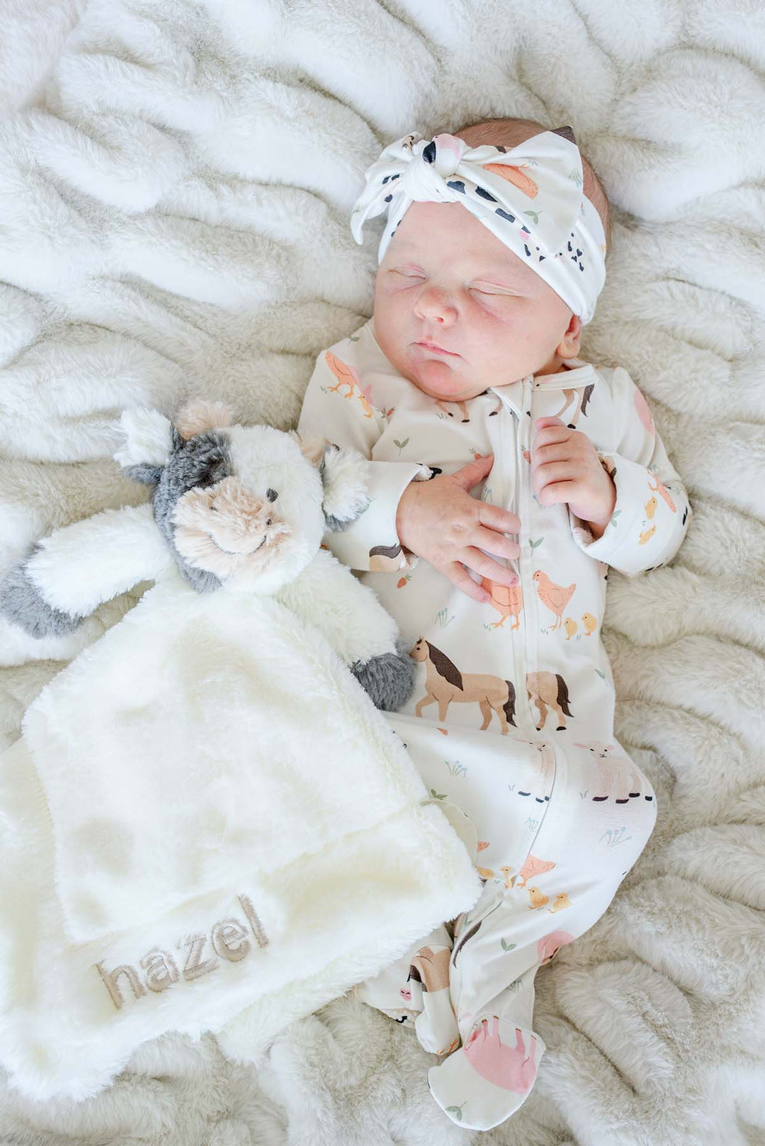 Newborn in a farm animal sleeper next to a cream and grey Caden Lane cow lovey embroidered "hazel"