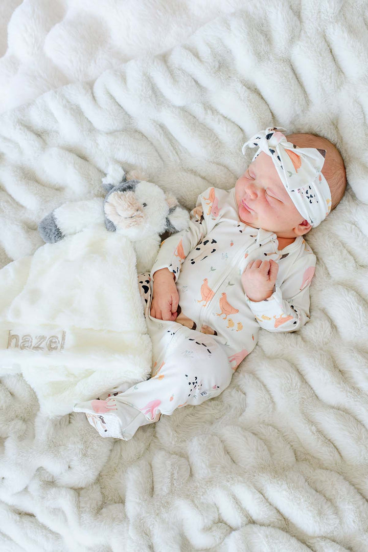 Newborn asleep on a white faux fur blanket beside a personalized Caden Lane cow lovey