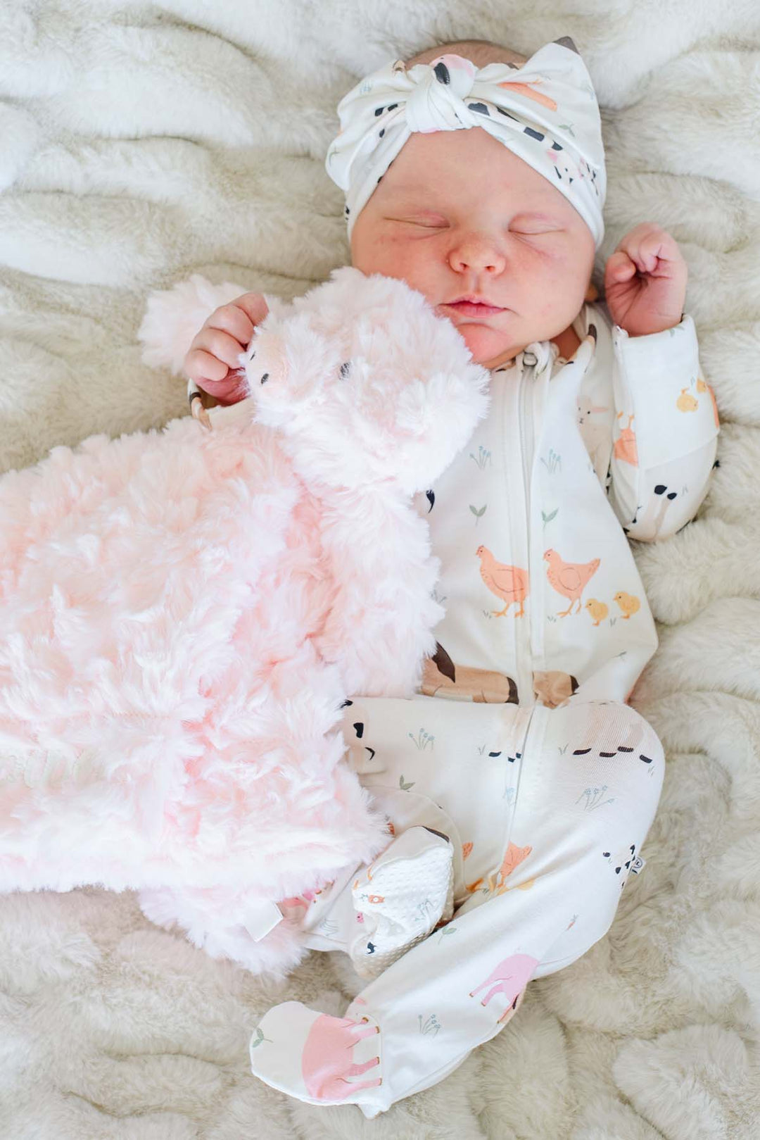 Sleeping newborn in a farm animal sleeper holding a soft pink Caden Lane pig lovey