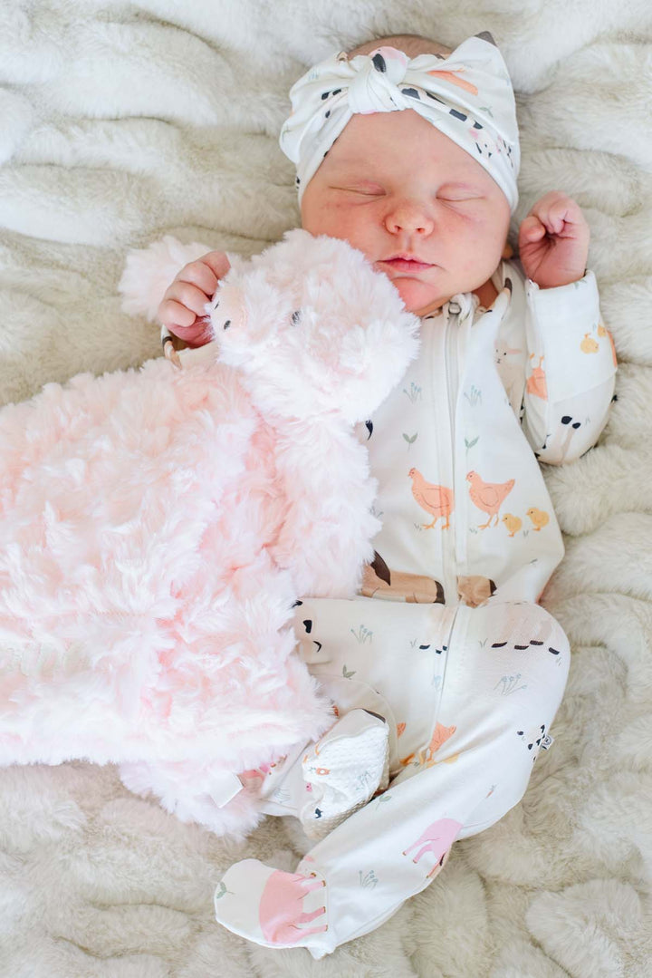 Sleeping newborn in a farm animal sleeper holding a soft pink Caden Lane pig lovey