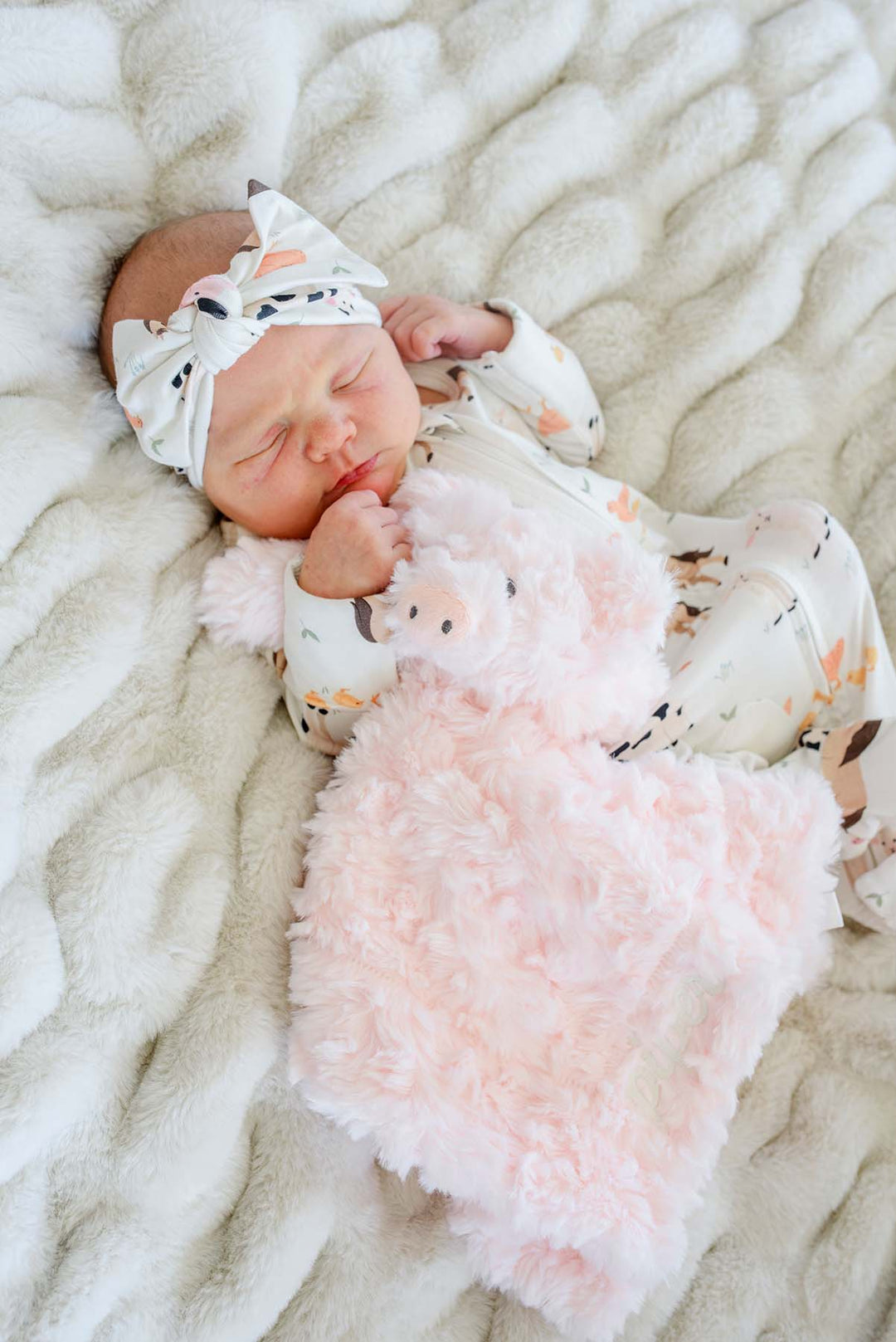 Newborn in a bow headband snuggled under a personalized Caden Lane pink pig lovey