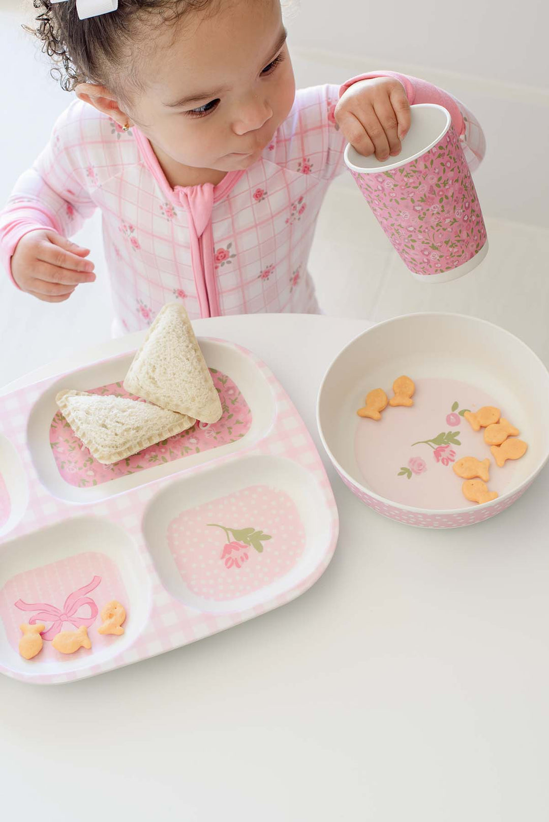 A young child in Caden Lane pink floral pajamas sits at a white table, holding a pink floral cup. In front of the child is a pink gingham divided meal tray with two paper cone snacks and a matching pink bowl with small orange snacks.