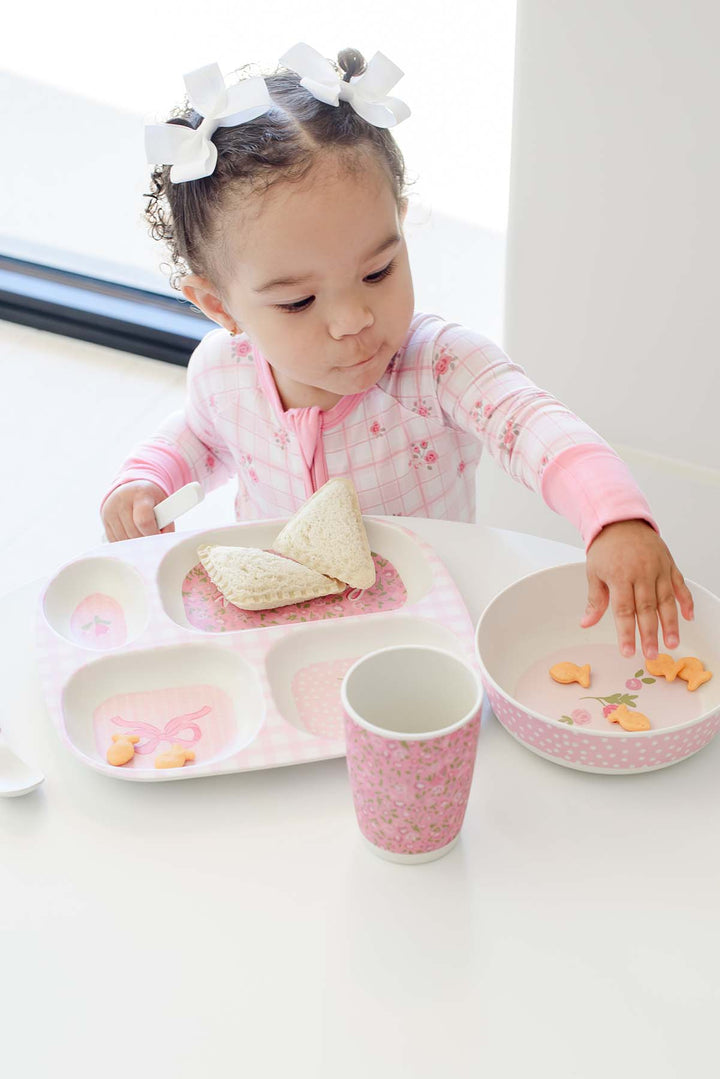 A toddler girl wearing a pink floral long-sleeve shirt and white bow headband sits at a white table, using a pink and white divided tray with a matching cup and bowl.