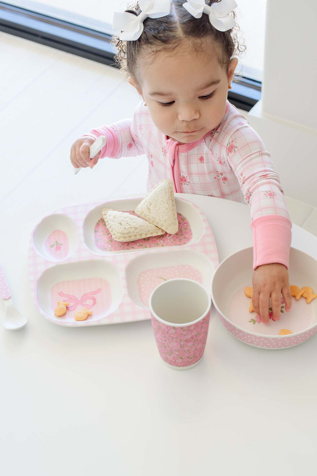 A toddler girl wearing a pink long-sleeve pajama top sits at a white table, using a pink and white butterfly-themed children's dinner set. The set includes a three-compartment tray with a butterfly design, a matching cup, and a bowl.
