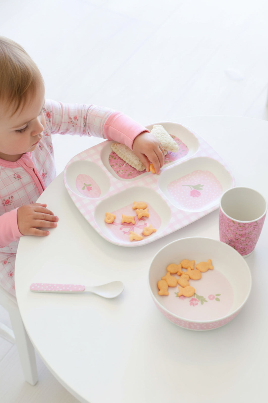 A child wearing a pink floral long-sleeve top uses a pink and white divided baby meal tray with a matching cup and bowl on a white table.