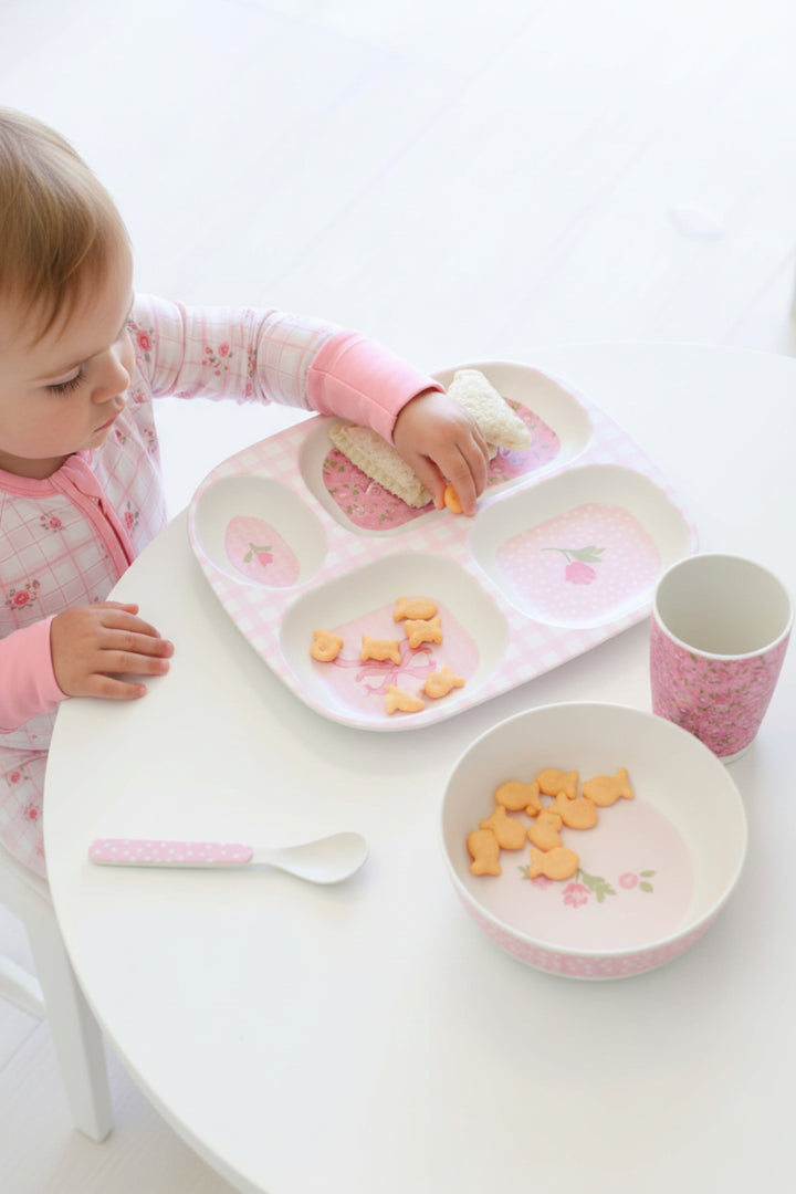 A child wearing a pink floral long-sleeve top uses a pink and white divided baby meal tray with a matching cup and bowl on a white table.
