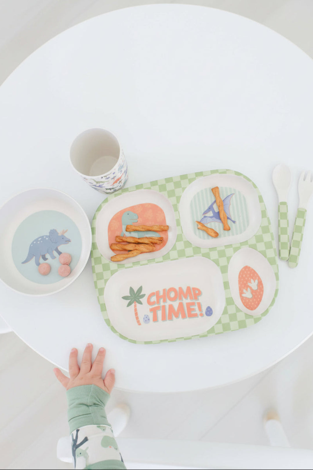 A children's dinner set arranged on a white table, including a green-and-white checkered four-compartment tray with the words 'CHOMP TIME!' and dinosaur-themed food, a matching bowl, a patterned mug, a fork, and a child's hand resting near the tray.