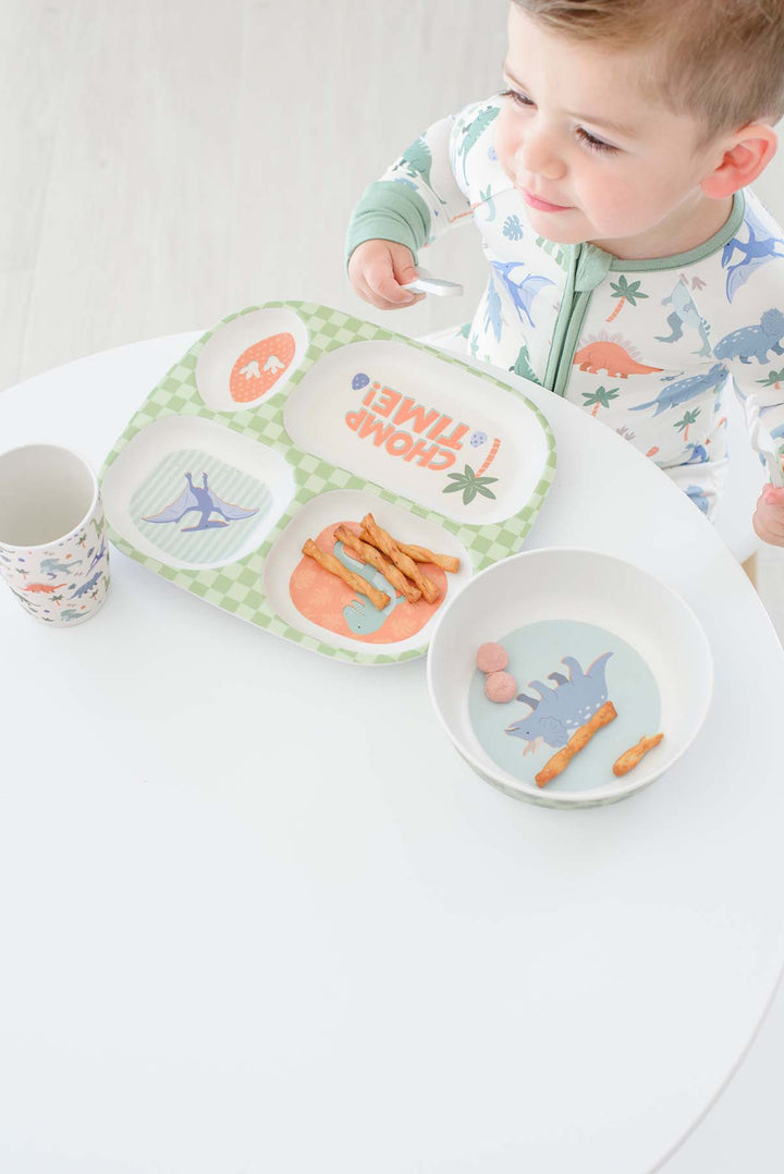 A toddler wearing dinosaur-print pajamas sits at a white table with a green and white divided meal tray, a small bowl, and a cup. The tray has colorful dinosaur illustrations and the words 'CHOMP TIME!'.