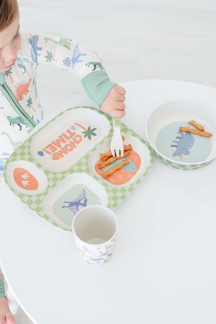 A child wearing a dinosaur-print pajama top sits at a white table using a green and white compartment tray with dinosaur illustrations. The tray has sections and includes matching children's tableware, including a cup and a bowl.