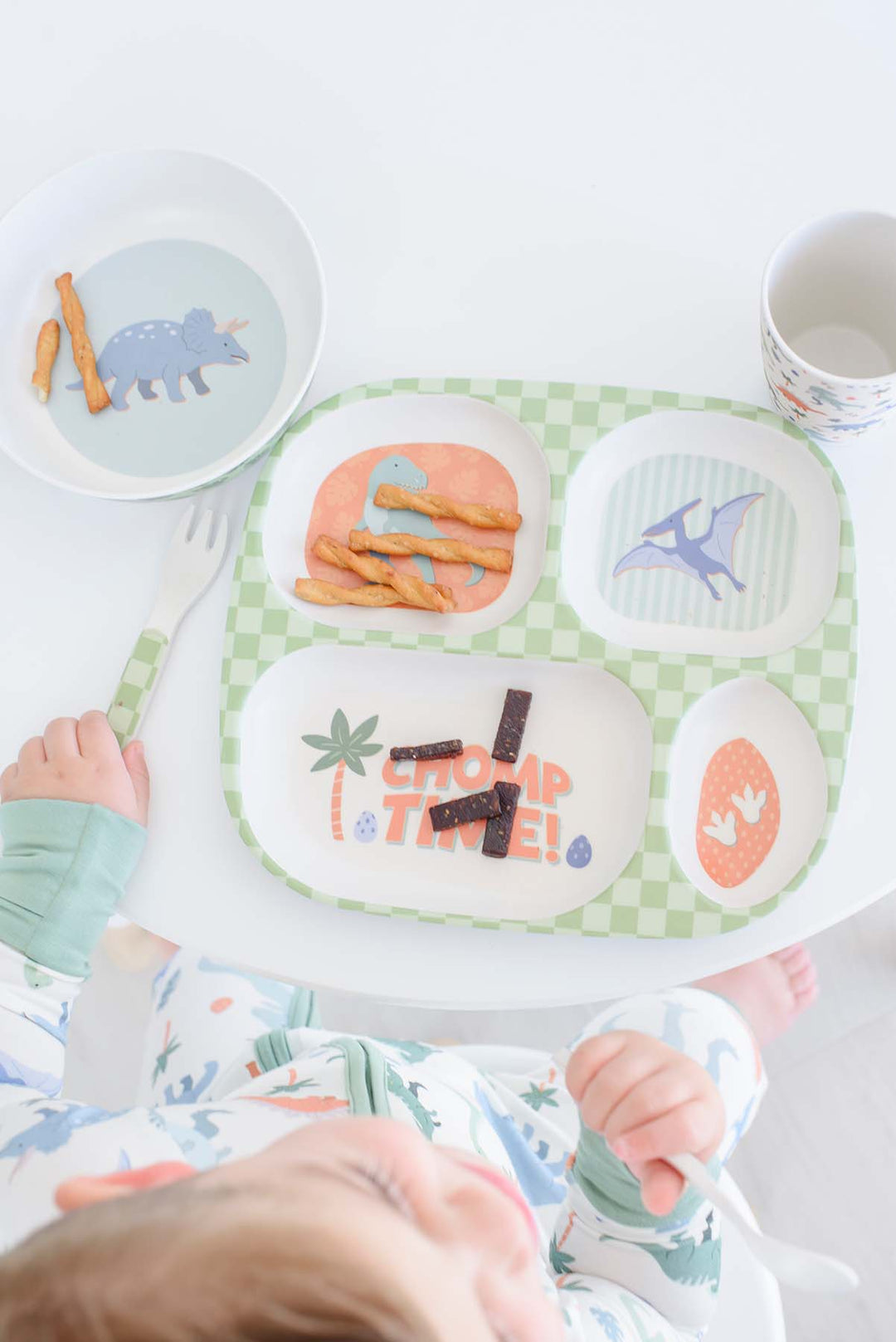 A baby in a patterned onesie sits at a white high chair table, looking down at a green and white checkered divided meal tray with dinosaur and palm tree illustrations. The tray contains snacks, next to a matching bowl and a white plastic fork.