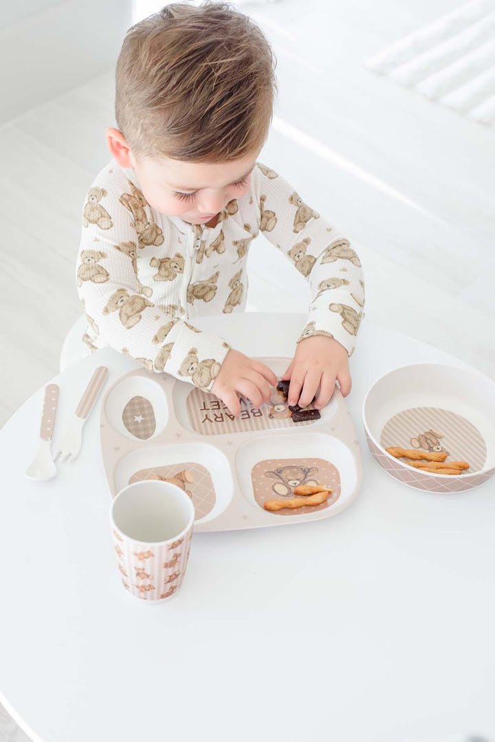 A toddler wearing a cCaden Lane cream long-sleeve pajama shirt with a brown teddy bear pattern sits at a white table, using a Caden Lane beige divided meal tray with a matching cup and bowl.