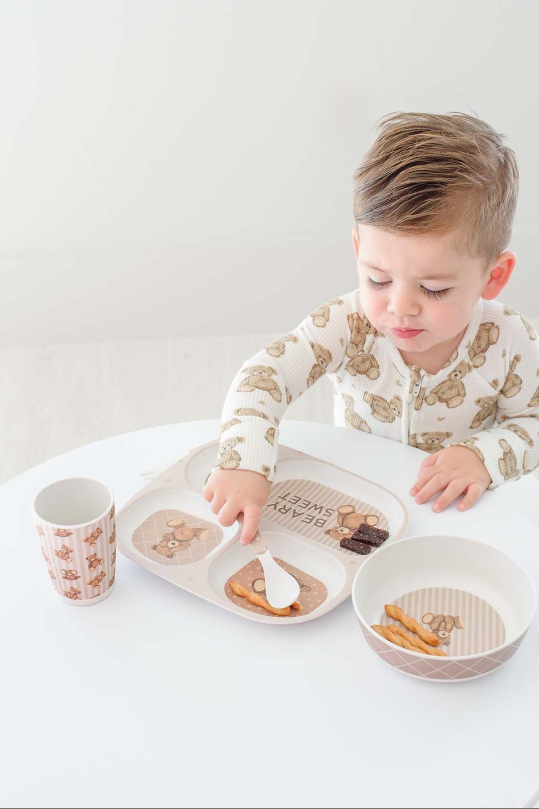 A young child wearing Caden Lane cream pajamas with a brown animal print sits at a white table, reaching for food on a divided tray. A matching Caden Lane patterned cup and bowl are also on the table.