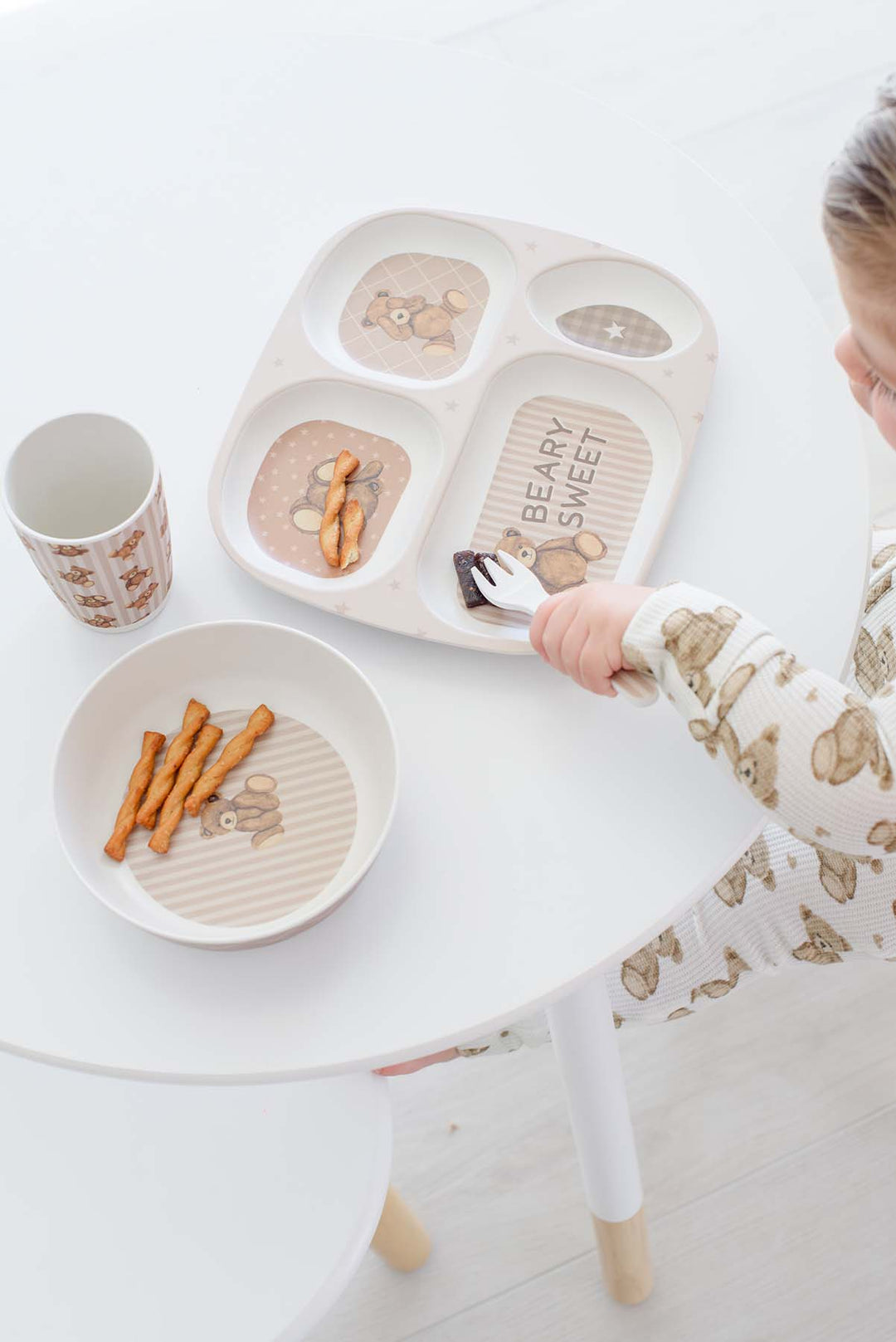 A child wearing Caden Lane beige bear pajamas sits at a white table using a Caden Lane beige compartment baby tray with food. The tray features cartoon bears and the words 'BEARY SWEET'.
