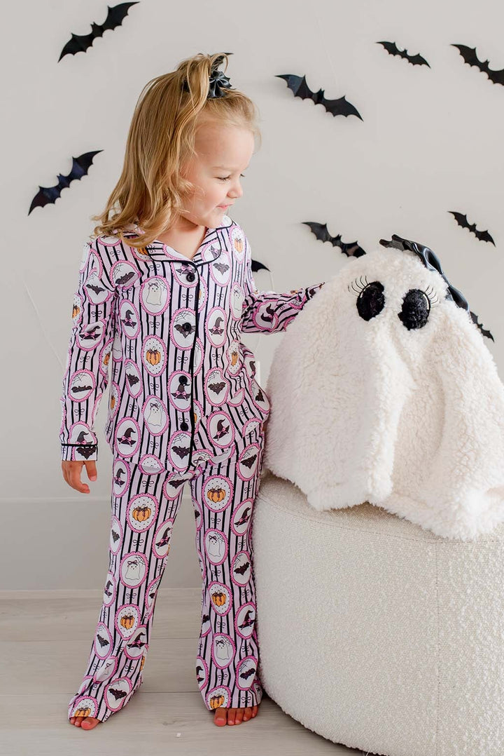 Girl standing next to plush ghost and wearing Caden Lane pink and black striped Halloween pajamas with ghost, bat, and pumpkin cameo print button-up two-piece set.