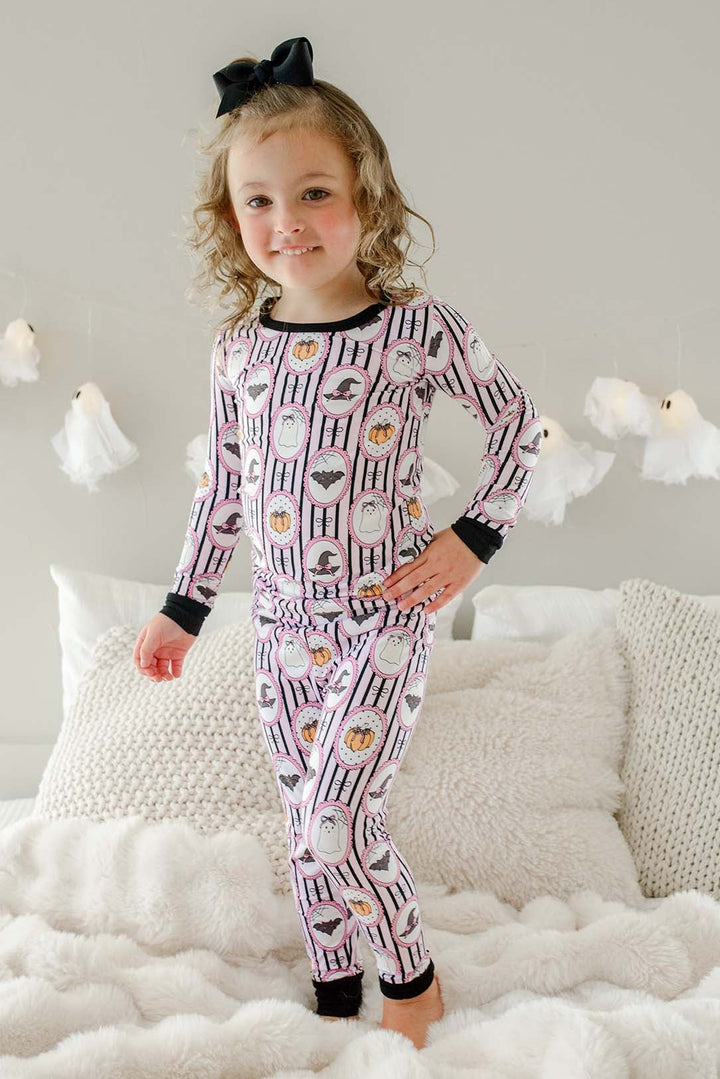 Young girl standing on a bed wearing a Caden Lane pink and black striped two-piece pajama set with ghost, bat, and pumpkin cameo print, with a black bow in her hair and hanging ghost lights in the background.