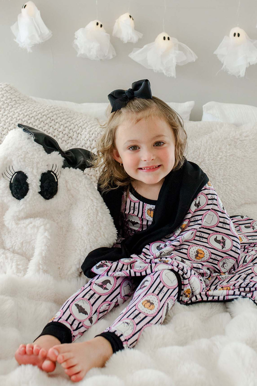 Child wrapped in a Caden Lane pink and black striped bamboo blanket with ghost, bat, pumpkin, and witch hat cameo print, standing on a couch with hanging ghost lights in the background
