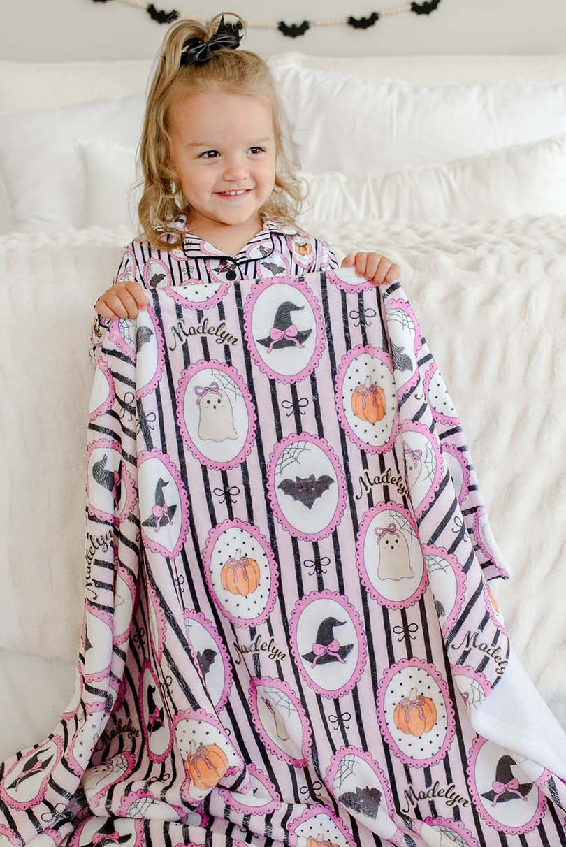 A young girl sitting on a white bed, holding up a pink and black patterned blanket with Halloween-themed illustrations like ghosts, pumpkins, bats, and witch hats.